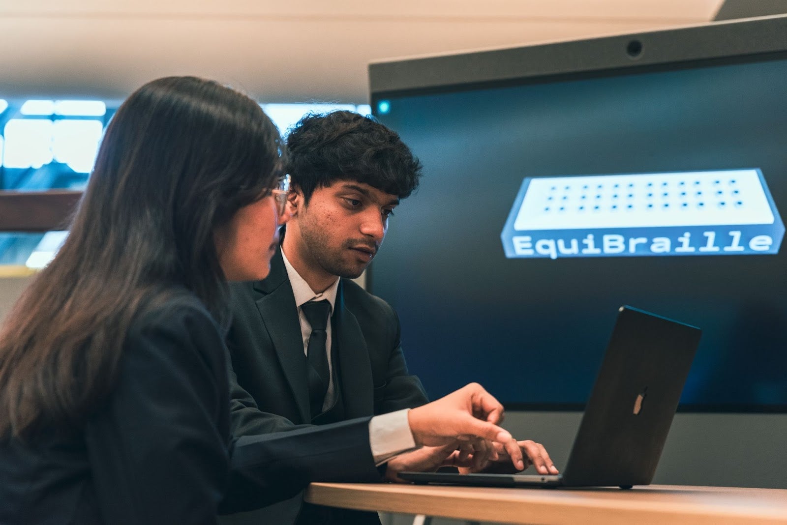 Two students work on a laptop while a screen behind them displays the text "EquiBraille" on a 3D render of the product.