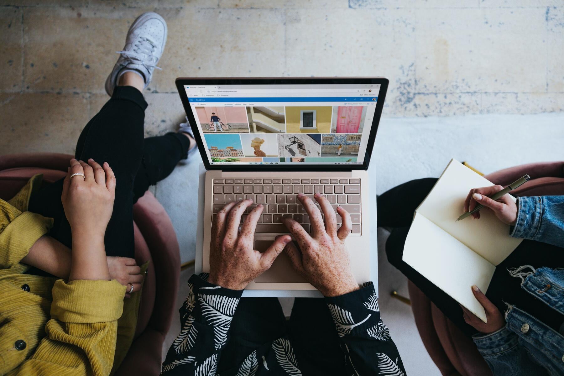 Three individuals are seated next to one another, sharing the same computer. One person sits to the side, taking notes as another types.