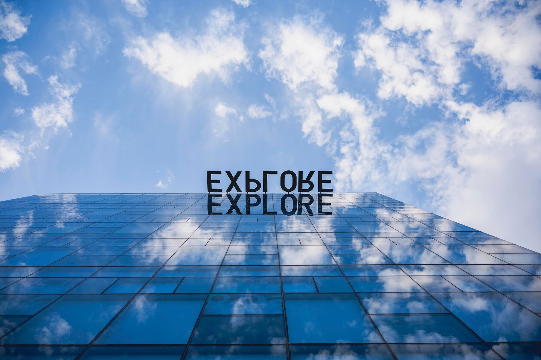 View of a building from the ground up with blue skies, white cloud and the word Explore written on the building reflection. 