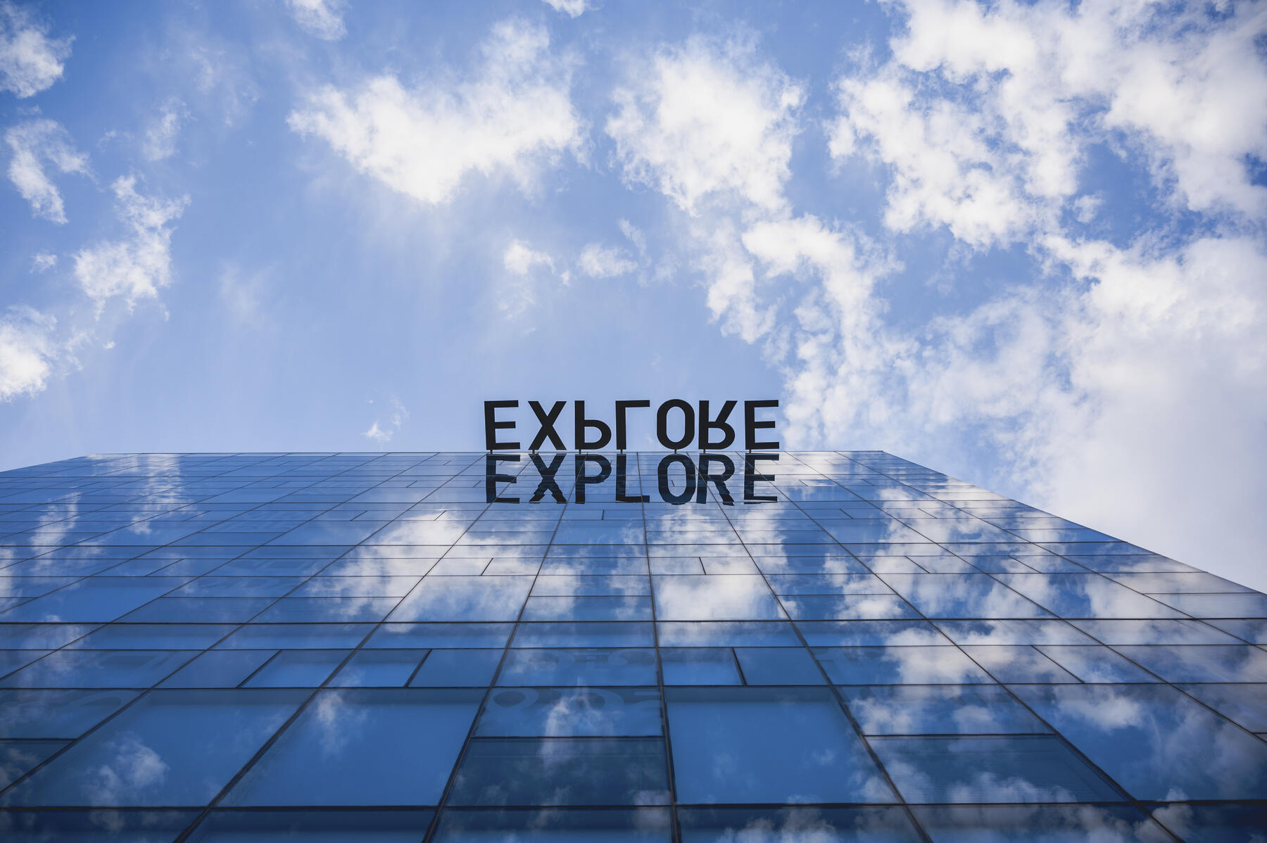 View of a building from the ground up with blue skies, white cloud and the word Explore written on the building reflection. 