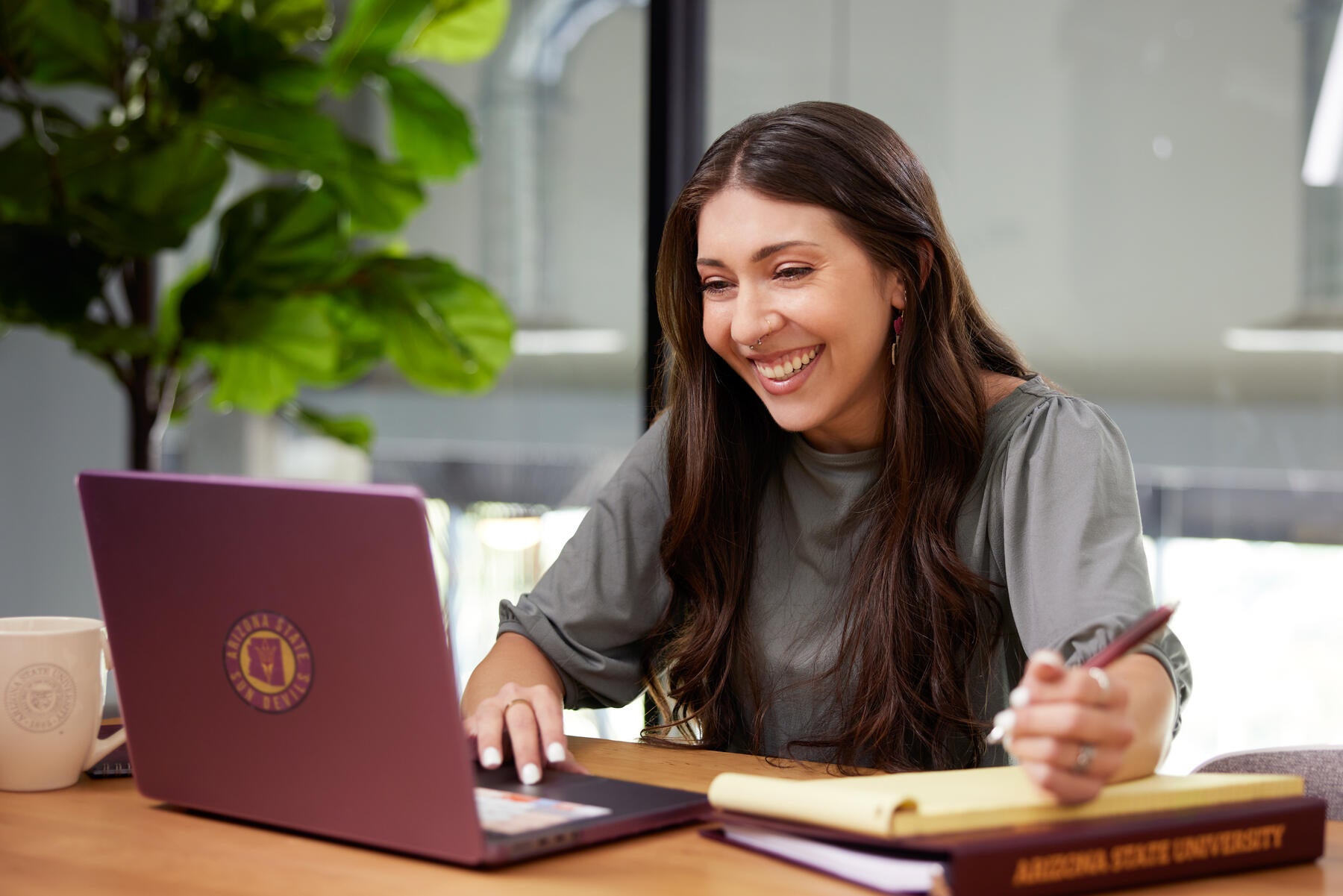 Person smiling and working on a laptop at a table with a notebook and Arizona State University folder.