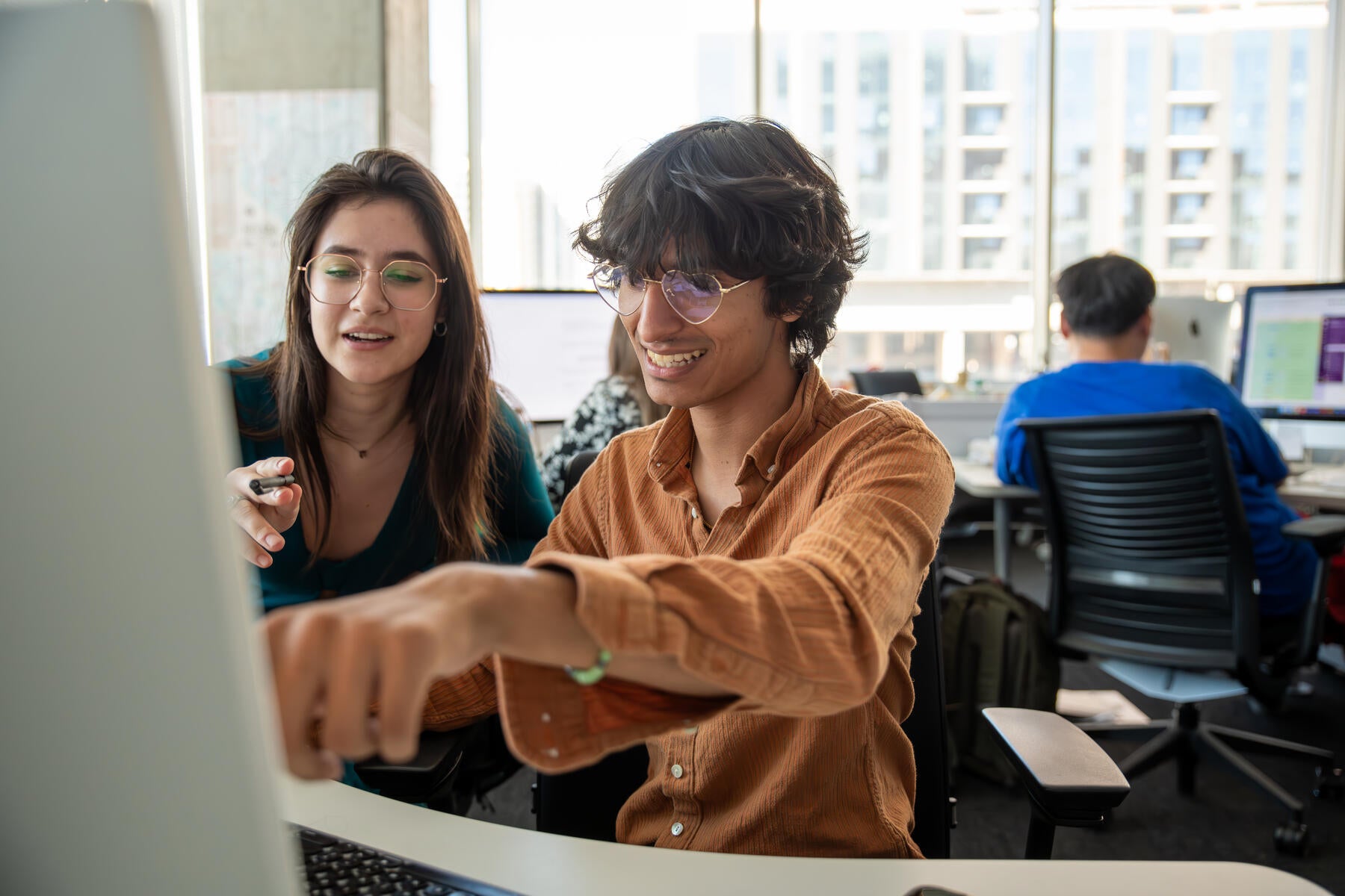 Two students behind a computer screen, with one in an orange shirt pointing at the screen