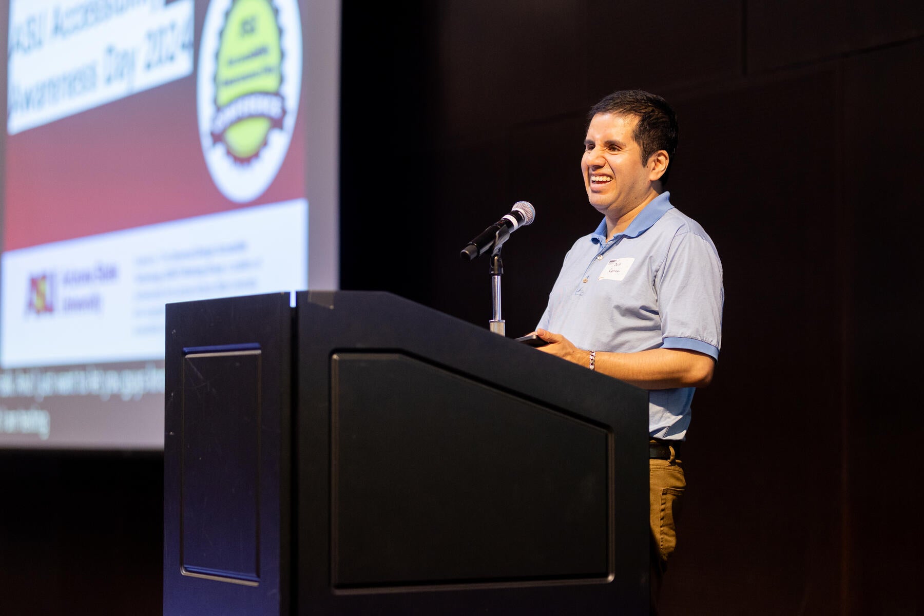 Man in stripped polo standing on stage behind a podium with screen to the left