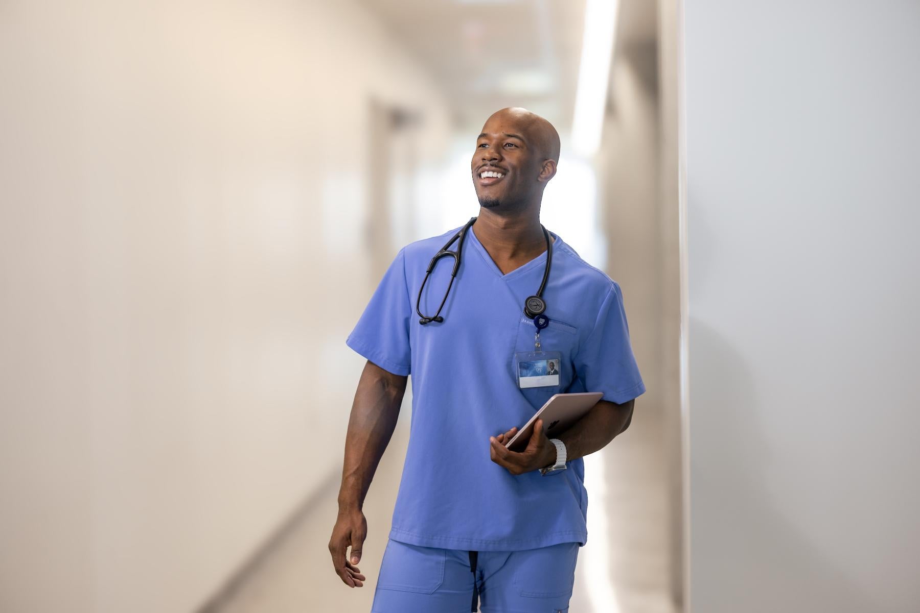 A man in blue nursing scrubs standing in a hallway with a laptop in his hand