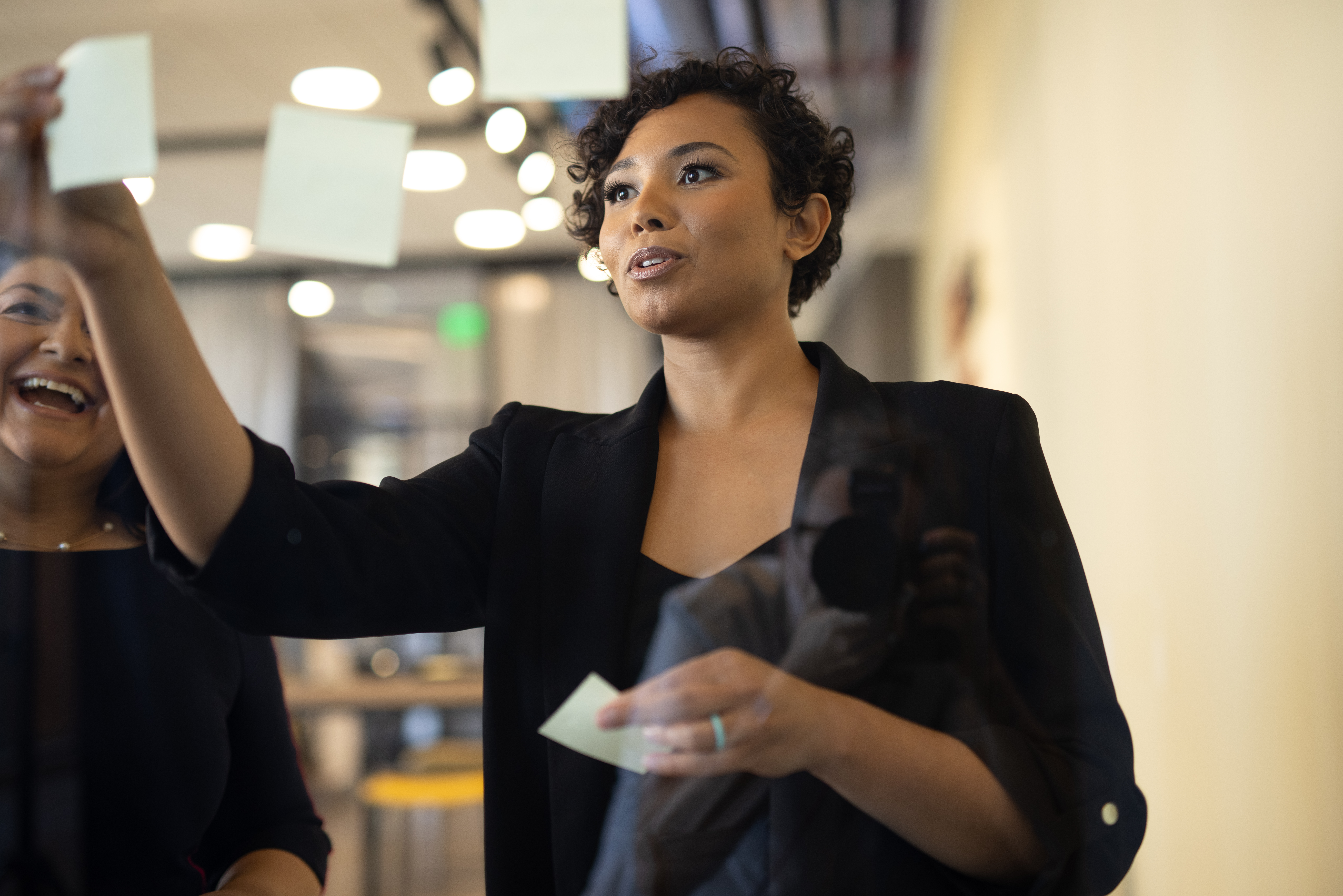 Two women place sticky notes on a glass wall, observing the notes they've jotted down with a smile.