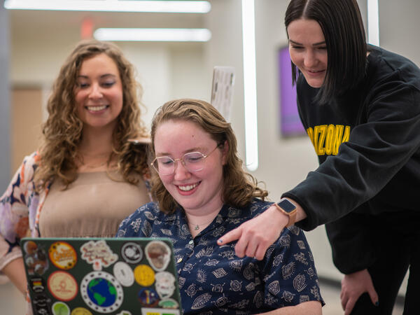 Three woman huddle around a laptop