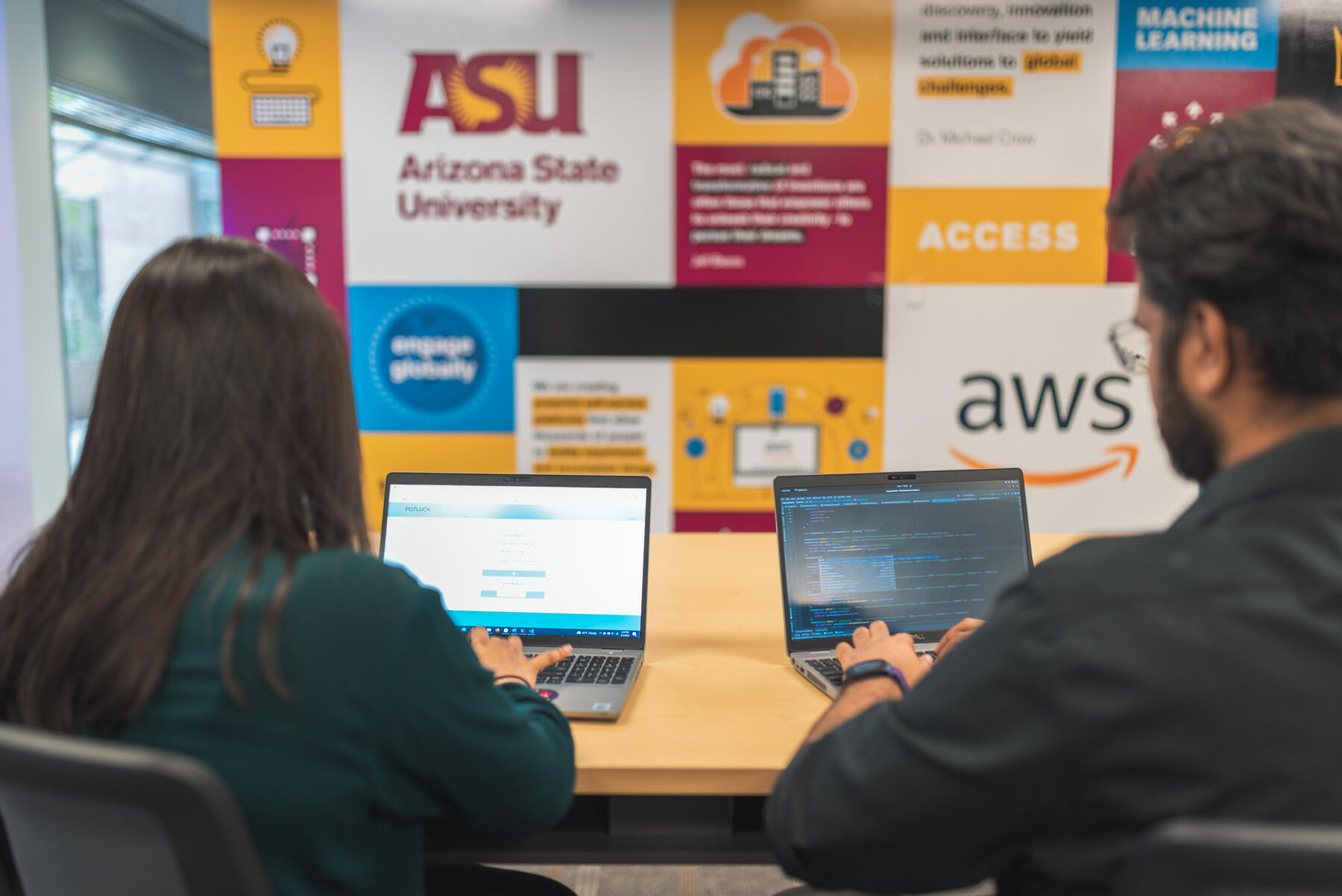 Two students sitting at a table with their laptops and backs to the camera