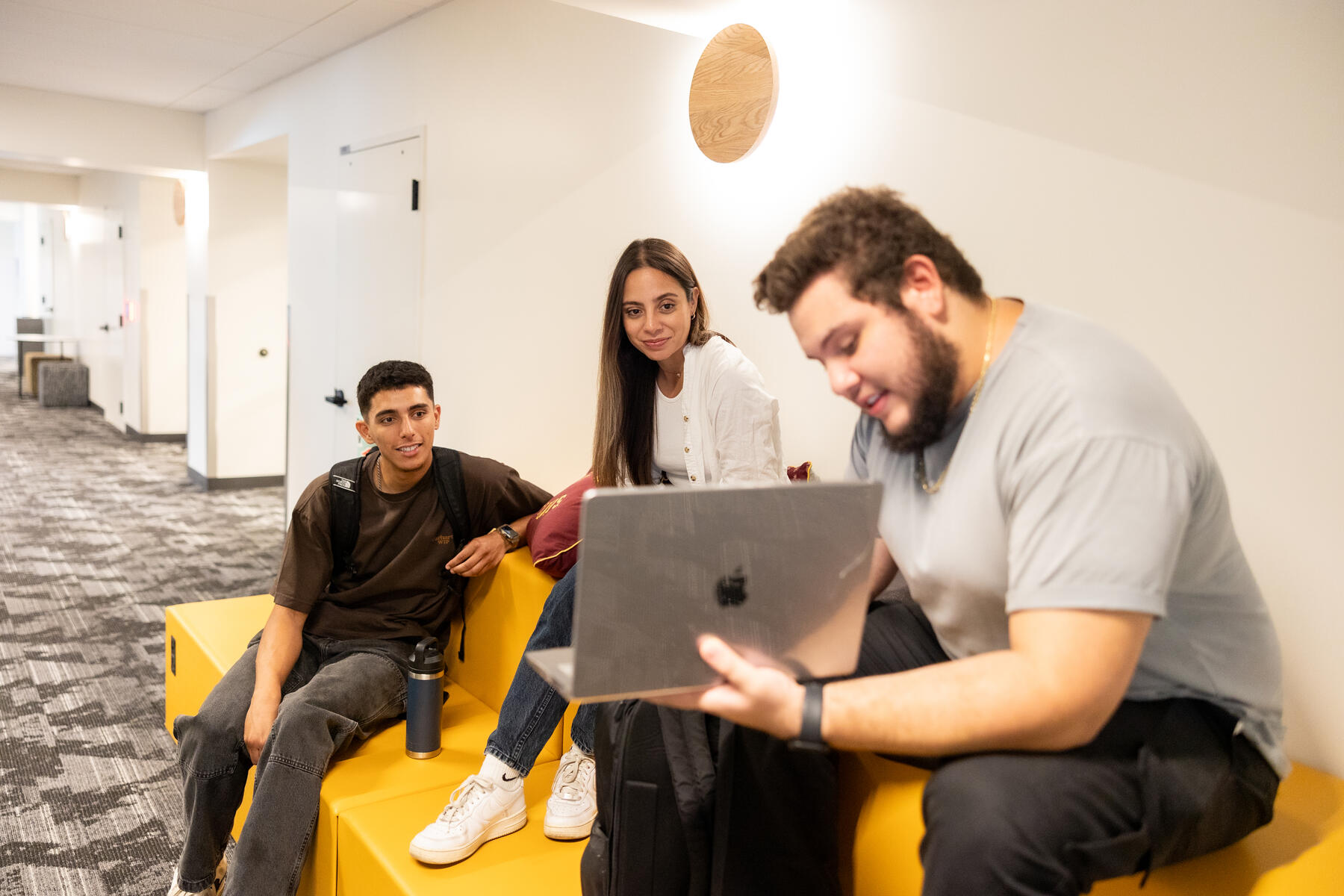 Three young adults sitting on yellow seating in a hallway, looking at a laptop.